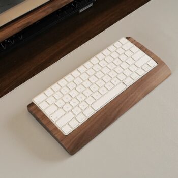 A top view of an Apple Magic Keyboard in a walnut wood tray, paired with a matching walnut wood tray holding a Magic Trackpad, placed on a desk with a monitor stand in the background. The trays, made from sustainably sourced walnut, create a unified workspace aesthetic.