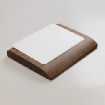 An angled side view of an Apple Magic Trackpad in a walnut wood wrist rest tray, highlighting the compact footprint and micro-suction base for desk stability. The tray, made from sustainably sourced walnut with a 12-month wood integrity guarantee, is displayed on a wooden surface.