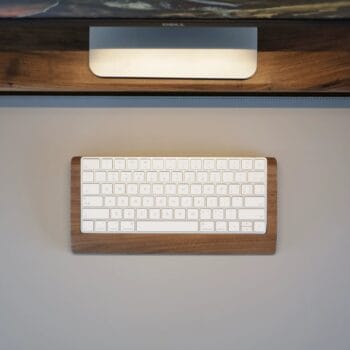 A top view of an Apple Magic Keyboard in a walnut wood tray, placed on a desk with a monitor stand in the background. The tray, crafted from FSC-certified walnut with a 24-month wood integrity guarantee, features a non-slip felt base.