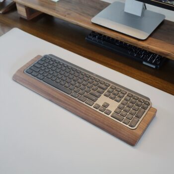 A top view of a Logitech Craft Keyboard in a walnut wood tray, placed on a wooden desk with a monitor stand in the background. The tray, designed for modular compatibility with Magic Keyboard trays, is made from sustainably sourced walnut.