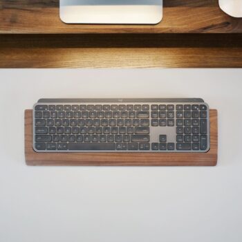 A top view of a Logitech Craft Keyboard in a walnut wood tray, highlighting the precision-cut fit and 3mm felt lining for keyboard protection. The tray, crafted from FSC-certified walnut, is placed on a wooden desk with a monitor stand in the background.